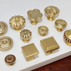 Op de afbeelding: Een verzameling van 12 gouden kastknoppen met verschillende ontwerpen, waaronder een bloem, een vierkant en een klavertjevier. De knoppen zijn gerangschikt op een wit oppervlak.