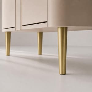 Puede incluir: Un mueble de color beige claro con un acabado mate liso y esquinas redondeadas. La pieza está sostenida por tres patas cilíndricas cónicas visibles, hechas de metal pulido en tono dorado, que proporcionan una base firme a la estructura.