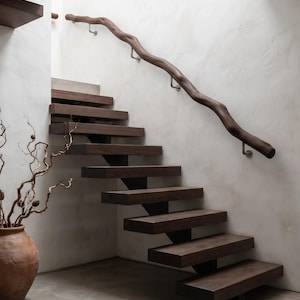 May include: A staircase featuring dark brown wooden floating steps and a matching organic, wavy wooden handrail mounted to a white wall with silver metal brackets. A large terracotta-colored ceramic vase with dried branches sits nearby.