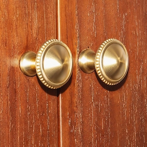Op de afbeelding: Twee gouden kastknoppen met een parelrand en een licht gewelfde midden.