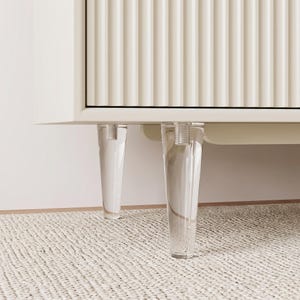 May include: A cream-coloured cabinet featuring a vertical fluted texture on the front panel. The furniture is supported by two transparent, conical acrylic legs that attach to the base with visible silver-toned threaded metal hardware.