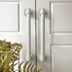 May include: Two white industrial pipe door handles mounted on a white door. The handles are made of metal pipe with a white finish. The door is a white paneled door.