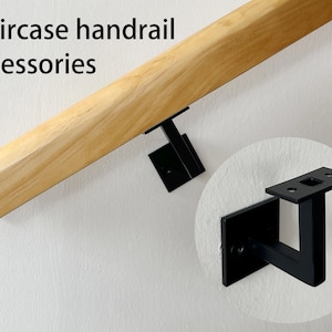 Puede incluir: Soporte de pasamanos de metal negro montado en una pared con un pasamanos de madera adjunto. El soporte tiene un agujero cuadrado en la parte superior para que el pasamanos pueda pasar.  "Staircase handrail accessories" está escrito en negro en la parte superior de la imagen.