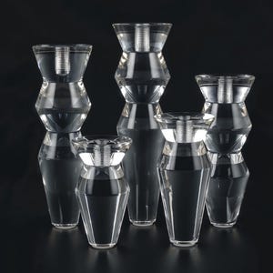 以下が含まれることがあります： 幾何学模様の透明なガラス製のキャンドルホルダーが5つ。キャンドルホルダーはサイズと形が異なり、すべて黒い表面に一列に並んでいます。