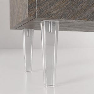 Puede incluir: Dos patas de mueble de acrílico transparente y cónicas fijadas a la parte inferior de un gabinete oscuro con vetas de madera. Cada pata presenta un perno roscado de metal plateado visible en la parte superior, que conecta el soporte transparente con la base de madera texturizada.