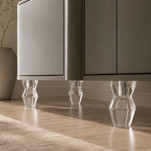 May include: A grey cabinet featuring three clear, faceted acrylic furniture legs with a geometric, tapered design. The legs are attached to the base of the cabinet, providing a transparent support structure that elevates the main unit from the floor.