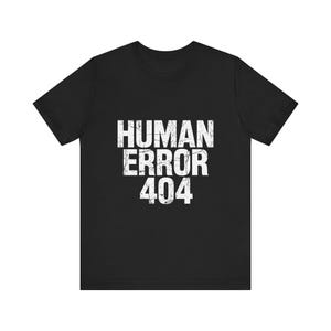 Op de afbeelding: Een zwart t-shirt met korte mouwen en de witte, verweerde tekst "HUMAN ERROR 404" in het midden op de borst. De afbeelding heeft een gebarsten, versleten uiterlijk en het shirt is voorzien van een standaard ronde hals en een rechte zoom.
