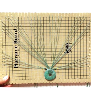May include: A wooden macrame board with a grid pattern and a turquoise bead in the center. The board is labeled "Macrame Board" and "designed by Anne Dिल्कर for the BeadSmith".