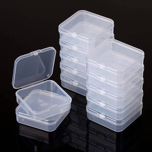 May include: A stack of clear, square plastic hinged-lid containers. One container is open, revealing the interior. The containers are made of translucent plastic and are stacked on a black surface. The containers are small and appear to be for storing small items.