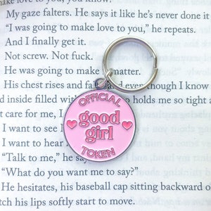 Może przedstawiać: Różowy metalowy brelok do kluczy z napisem "Official Good Girl Token" i dwoma różowymi sercami. Brelok jest przymocowany do srebrnego kółka na klucze.