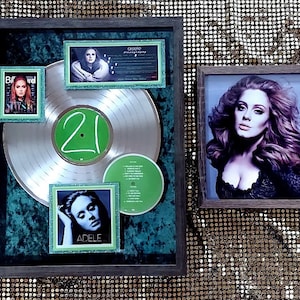Puede incluir: Un disco de platino enmarcado con un fondo verde y un disco plateado con el número "21" en verde. Hay una foto enmarcada más pequeña de Adele a la izquierda y una foto enmarcada más grande de Adele a la derecha.