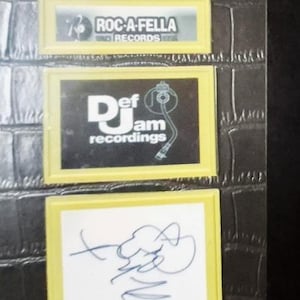 May include: Close-up of a black surface with three rectangular yellow-framed images. The top image displays "ROC-A-FELLA RECORDS". The middle image shows "Def Jam recordings". The bottom image features a blue signature on a white background.