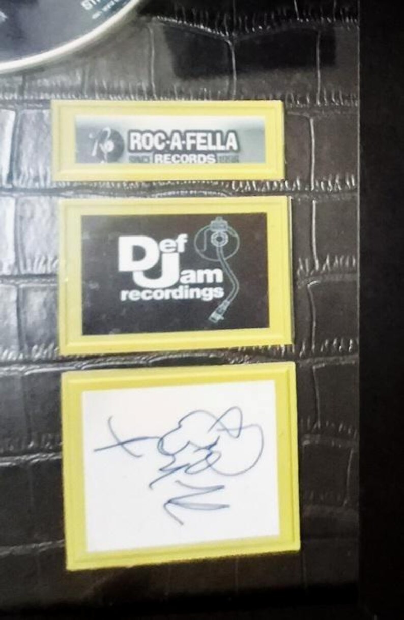 May include: Close-up of a black surface with three rectangular yellow-framed images. The top image displays "ROC-A-FELLA RECORDS". The middle image shows "Def Jam recordings". The bottom image features a blue signature on a white background.