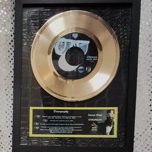 May include: Framed gold record of Kanye West's "Stronger" single. The record is mounted on a black textured background within a black frame. A small plaque with discography details and a photo of Kanye West is included.