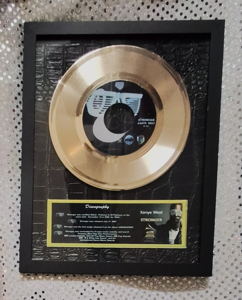 May include: Framed gold record of Kanye West's "Stronger" single. The record is mounted on a black textured background within a black frame. A small plaque with discography details and a photo of Kanye West is included.