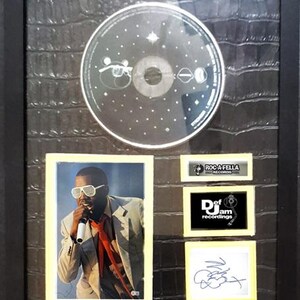 May include: Framed music memorabilia featuring a CD, a photograph of a performer wearing sunglasses, and a signature. The frame is black, and the background has a textured, dark pattern. Includes logos for Roc-A-Fella Records and Def Jam Recordings.