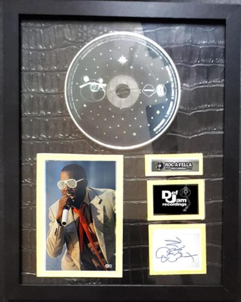 May include: Framed music memorabilia featuring a CD, a photograph of a performer wearing sunglasses, and a signature. The frame is black, and the background has a textured, dark pattern. Includes logos for Roc-A-Fella Records and Def Jam Recordings.