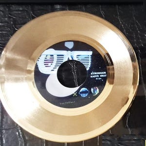 May include: A gold-colored vinyl record with a black and blue label. The label features the text "STRONGER KANYE WEST 45 rpm" and a graphic design. The record is a circular shape.