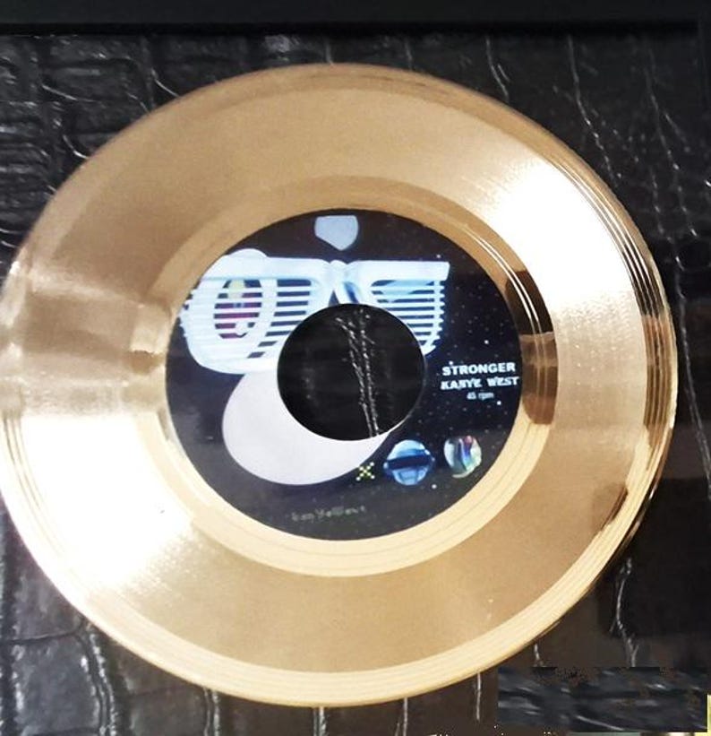 May include: A gold-colored vinyl record with a black and blue label. The label features the text "STRONGER KANYE WEST 45 rpm" and a graphic design. The record is a circular shape.