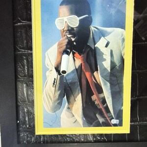 May include: A framed photograph of a person wearing white shutter shades and a red tie, holding a microphone. The person is wearing a light-colored suit jacket. The photograph is set against a blue background.