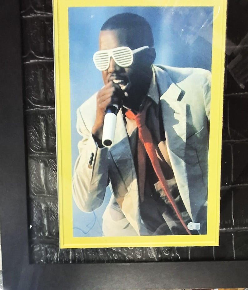 May include: A framed photograph of a person wearing white shutter shades and a red tie, holding a microphone. The person is wearing a light-colored suit jacket. The photograph is set against a blue background.