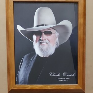 May include: A framed black and white photo of Charlie Daniels, a country music singer, wearing a white cowboy hat and sunglasses. The photo is in a wooden frame with a black mat. The text below the photo reads "Charlie Daniels, October 28, 1936, July 6, 2020."