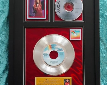 Könnte beinhalten: Eine gerahmte Platin-Schallplatte für Reba McEntires Song "Fancy". Der Rahmen enthält ein Foto von Reba McEntire, eine CD und eine kleine Plakette mit Text über den Song. Der Hintergrund ist rot.