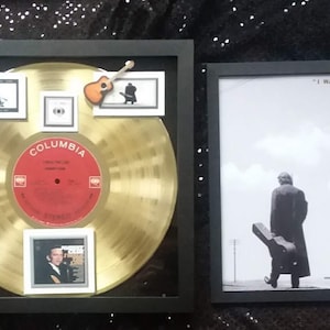 Op de afbeelding: Een ingelijste gouden plaat met een zwarte passe-partout en een rood label met de tekst "Columbia" en "I Walk the Line" van Johnny Cash. De plaat is omringd door kleinere foto's en een miniatuur-gitaar. De lijst staat naast een zwart ingelijste foto van Johnny Cash die met een gitaarkoffer in zijn hand van de camera wegloopt. De foto is ondertiteld "I Walk the Line - Johnny Cash 1932-2003".