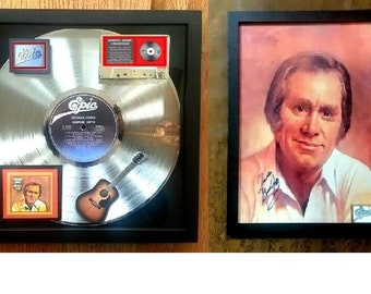 May include: A framed silver record award with a miniature guitar and a cassette tape. The record is for George Jones's album "Greatest Hits". The frame also includes a smaller photo of George Jones with his signature. The photo is framed in black.