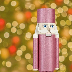 Può includere: Una figura di schiaccianoci con un cappello cilindrico e un cappotto lungo, entrambi ricoperti da una finitura glitterata rosa scintillante. La figura ha un viso pallido, occhi blu, baffi bianchi e una bocca rettangolare con denti bianchi.
