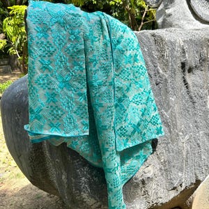 May include: A turquoise textile with an embroidered pattern. The fabric has a repeating geometric and floral design. The textile is draped over a stone sculpture, with a blurred green background of foliage. The textile is a vibrant shade of turquoise.