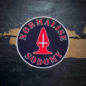 May include: A circular sticker with a dark blue background and white border. The sticker features the words "NORMALISE SODOMY" in red, arched around a red graphic. The graphic is a stylized, red, plug-shaped design with a white cross.