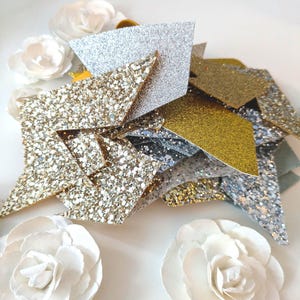 Puede incluir: Decoraciones en forma de estrella con purpurina dorada y plateada, junto con flores de papel blanco. Las estrellas tienen una superficie texturizada y brillante. El tema general es festivo y decorativo.