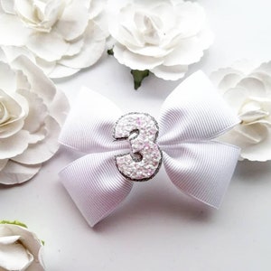 May include: White satin bow with a silver glitter number 3 in the centre. The bow is surrounded by white paper flowers.