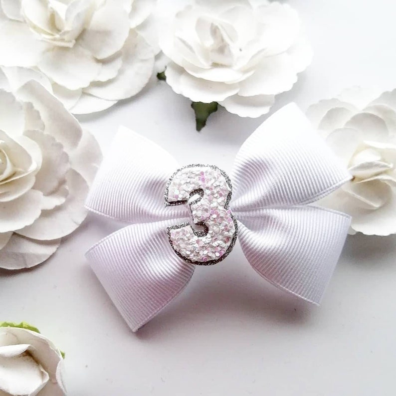 May include: White satin bow with a silver glitter number 3 in the centre. The bow is surrounded by white paper flowers.