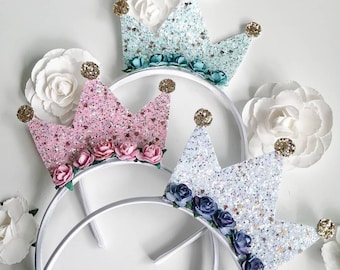 Puede incluir: Tres coronas de princesa de purpurina con diademas blancas y adornos florales. Las coronas son de color rosa, azul y blanco.