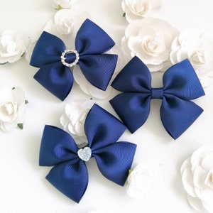 May include: Three navy blue hair bows with different embellishments. The first bow has a clear rhinestone ring in the centre. The second bow has a clear rhinestone heart in the centre. The third bow has a clear rhinestone ring in the centre.
