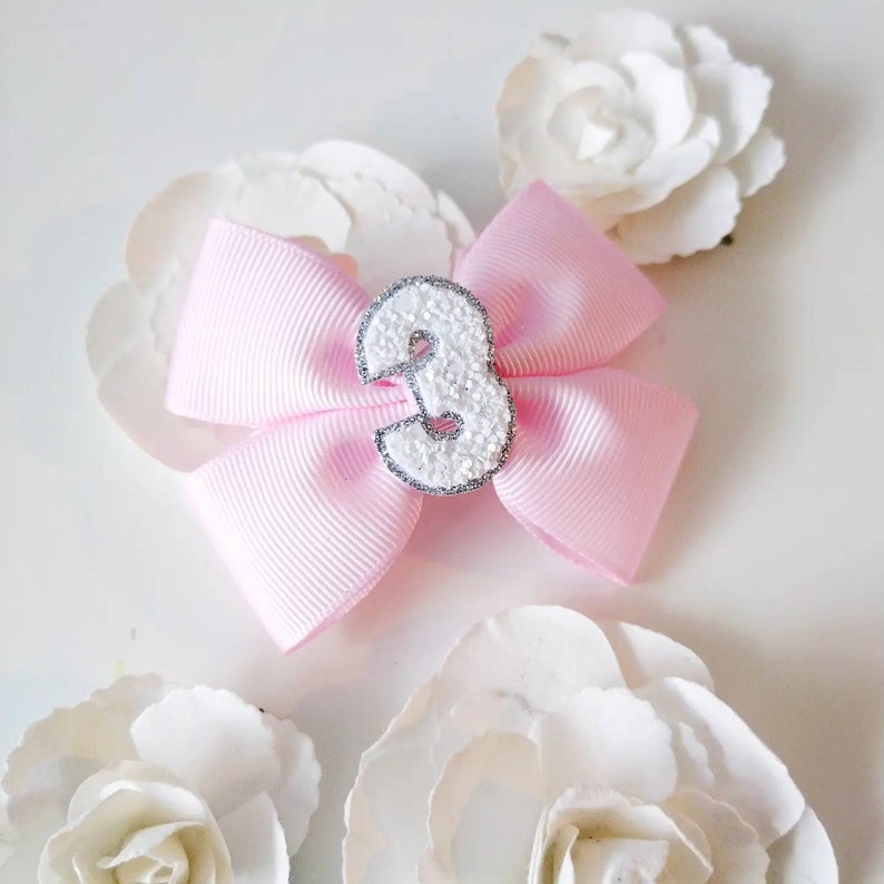 May include: A pink bow with a white glitter number three in the centre. The bow is surrounded by white flowers.