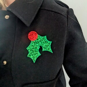 Puede incluir: Broche navideño festivo con una baya roja brillante y hojas de acebo verdes brillantes. La broche está sujeta a un abrigo negro, un accesorio de temporada.