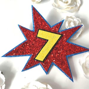 May include: A star-shaped cutout featuring a red glittery base with a blue glittery border. Centered on the red star is a yellow glittery number seven with a thin black outline. The design mimics a comic book style action burst graphic.