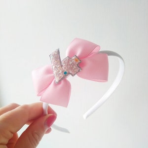 Könnte beinhalten: Ein rosa Stirnband mit einer weißen Schleife und einem glitzernden silbernen Buchstaben "K" in der Mitte. Das Stirnband ist perfekt für eine kleine Geburtstagsparty.
