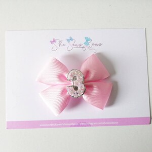 May include: Pink satin bow with a sparkly silver number 3 in the centre. The bow is attached to a hair clip.