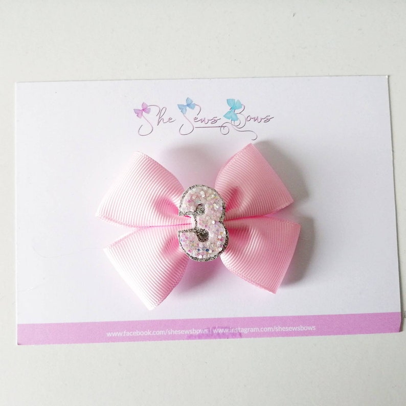 May include: Pink satin bow with a sparkly silver number 3 in the centre. The bow is attached to a hair clip.