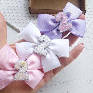 May include: Three hair bows with a sparkly number two on each. The bows are in pink, white, and lavender.