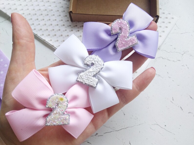 May include: Three hair bows with a sparkly number two on each. The bows are in pink, white, and lavender.