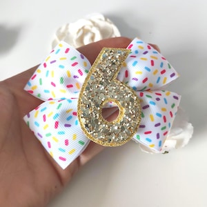 Könnte beinhalten: Eine weiße Schleife mit einem bunten Streumuster und einer goldenen Glitzerzahl sechs. Die Schleife ist ein Haarschmuck, ideal für eine Geburtstagsfeier. Die Schleife ist etwa 13 cm breit.