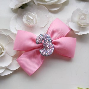 May include: A pink bow with a sparkly silver number 3 in the centre. The bow is on a white background with white flowers.
