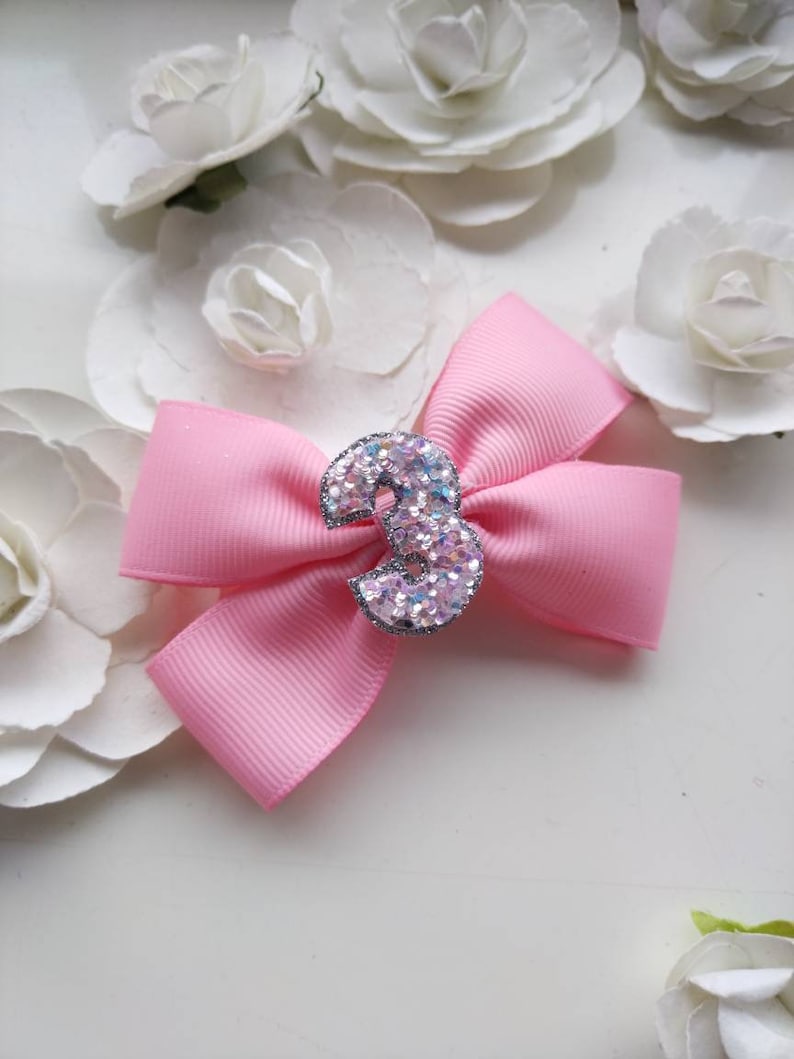 May include: A pink bow with a sparkly silver number 3 in the centre. The bow is on a white background with white flowers.