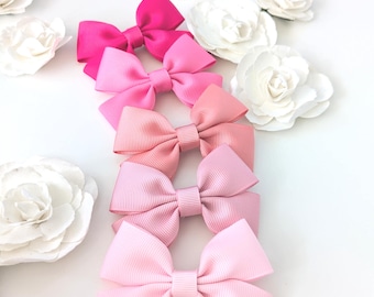 May include: Five pink ribbon hair bows in a gradient of shades, from vibrant fuchsia to a soft pastel pink. The bows are displayed on a white surface, with white paper flowers adding a delicate touch.