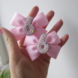 May include: Two pink hair bows with silver glitter number sixes in the centre. The bows are made of satin ribbon and have a sparkly finish.